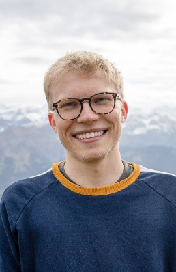 Photo de Janosch Herzog. M. Herzog porte des lunettes et sourit à la caméra. On ne voit que le haut de son corps. Il porte un pull-over bleu. En arrière-plan, on voit des montagnes et un ciel nuageux.